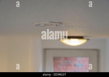 A view of a ceiling with an empty smoke detector and a light fixture. The smoke detector is white and circular, mounted on a textured ceiling. The lig Stock Photo
