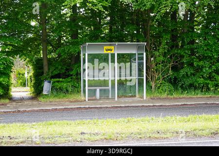 A small glass bus stop with a metal base and bench stands near a road, with a trash bin nearby and trees in the background. Stock Photo