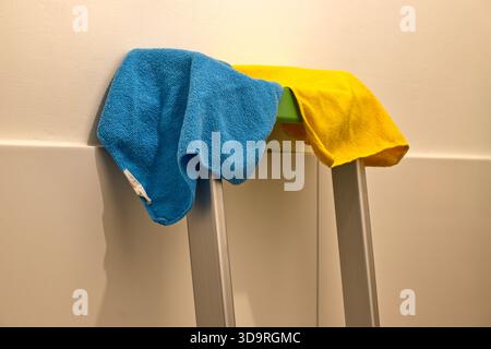 Two colorful microfiber cloths, one blue and one yellow, draped over a metal ladder against a plain wall. Stock Photo