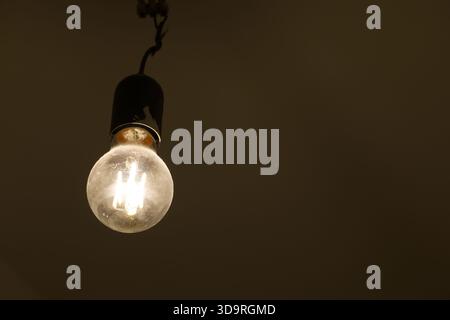 A single illuminated LED light bulb hanging from a ceiling in a dimly lit room. Stock Photo