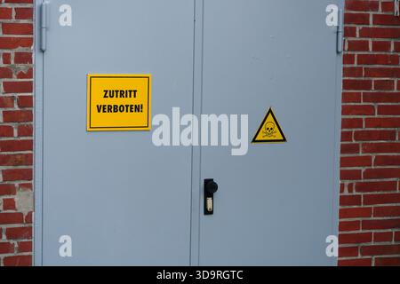 A red building features gray metal doors marked with yellow Zutritt verboten signs. Another door displays a triangular warning sign with a skull and c Stock Photo