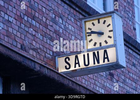 A red brick building features a large clock mounted on its facade. Below the clock hangs a white sign with the word Sauna clearly visible. The simple Stock Photo