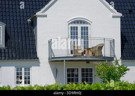Two story white house with a small metal balcony on the second floor. A round wooden table and two wicker chairs stand outside. Bushes nearby. Stock Photo