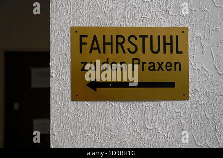 Gold colored sign mounted with four screws on uneven white painted wall showing German text Fahrstuhl zu den Praxen with left pointing arrow Stock Photo