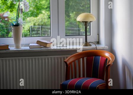 A striped blue and red vintage chair sits near a window with a marble sill. Three books, a white orchid in a pot, and an old lamp rest on the ledge. Stock Photo