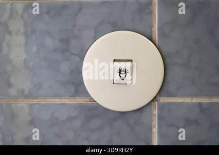 Old white colored light switch with bulb symbol mounted on blue tiles controlling upper lighting in room Stock Photo