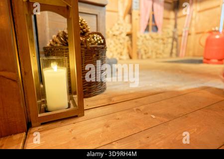 A white candle burns inside a wooden holder on a wooden floor. Behind it sits a woven basket filled with pinecones, with firewood and a window in the Stock Photo