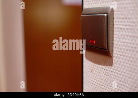 A silver switch with a glowing red indicator is mounted on a white wall. In the background, a brown door is visible. The indicator shows the on or off Stock Photo