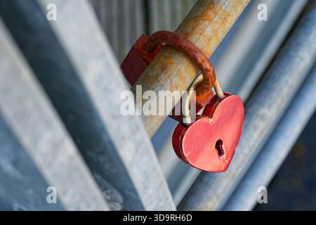 Two padlocks hang on a metal bar, one shaped like a red heart and the other heavily rusted. Both are covered in orange rust, symbolizing age and lasti Stock Photo