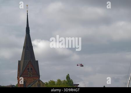 A tall historic church tower with a clock and rooster weather vane stands proudly while a red and white rescue helicopter flies by in the clear summer Stock Photo