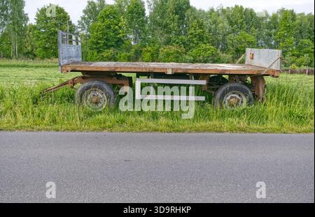 An old wooden trailer with rusty metal parts stands empty in a grassy field. Trees are visible in the background, suggesting a quiet rural setting wit Stock Photo
