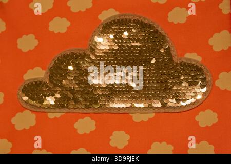 A white cloud shaped patch adorned with shiny silver sequins is sewn onto a red fabric background. The sparkling round sequins catch the light, symbol Stock Photo