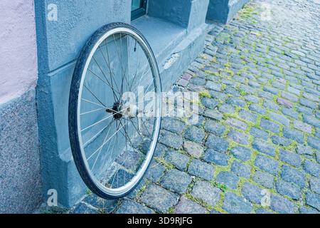 A single bicycle wheel is leaning against a gray building near a paved street. The urban scene shows worn cobblestones and the isolated wheel resting Stock Photo