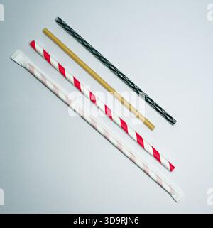On a white background lie four drinking straws made of paper, plastic and cardboard, and one black and green striped plastic straw. Stock Photo