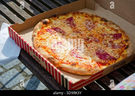 On a wooden bench lies an open pizza box labeled in German with the word Pizza. Inside is a delicious pizza topped with melted cheese and salami. Stock Photo