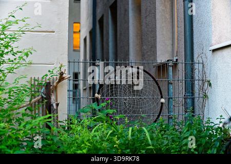 A small metal fence between two houses holds a black circular frame with a delicate woven floral like pattern in white. The design could be decorative Stock Photo