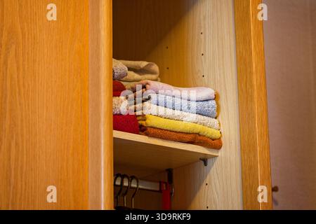 A wooden cabinet holds neatly folded towels in brown yellow white blue and pink arranged on shelves with larger towels beside them and hangers visible Stock Photo