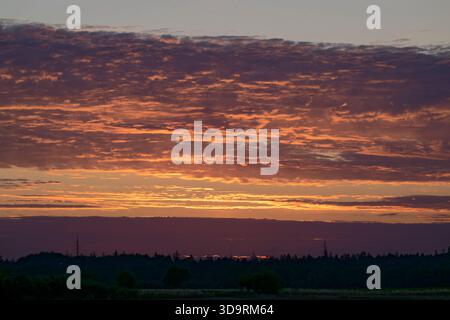 After sunset the sky glows in gold and orange, with layered clouds tinged in violet and peach. The low horizon shows a field with scattered trees, and Stock Photo