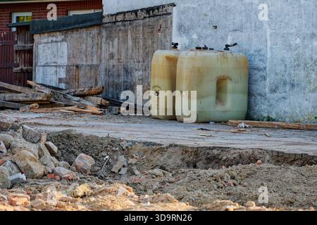 Two large canisters stand by a concrete wall in a torn up area. Broken bricks, soil and mud mix across the ground. An old tree and rough edges suggest Stock Photo