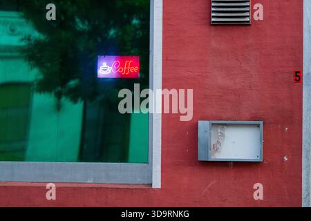 Behind glass in a red painted storefront window a bright neon sign shows a steaming coffee cup. The warm glow promises a cozy break and draws attentio Stock Photo