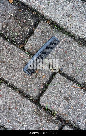 On pale paving tiles lies a small black comb, likely dropped or forgotten. Plastic teeth catch faint light, tiny scuffs show use. The quiet sidewalk s Stock Photo