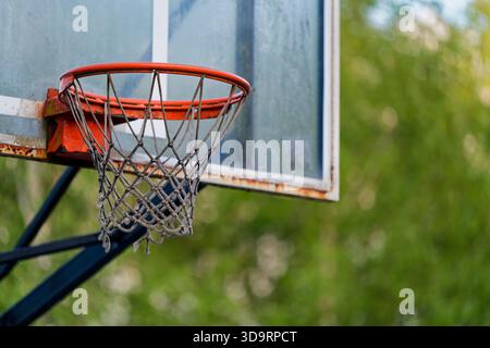 A tired court sits in greenery, a red hoop is rusted, the mesh net ripped and hanging in strands. Moss clings to metal and posts, dirt marks the frame Stock Photo