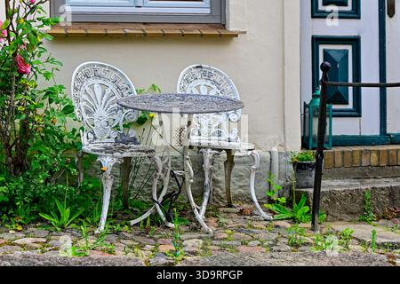 An old metal table and two delicate chairs rest beside a house entrance. White paint has cracked and flaked, gray metal shows through. A nearby rose s Stock Photo