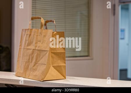 A plain paper bag with sturdy handles rests on a tidy gray shelf, light softly touches the creased sides, suggesting recent errands and simple storage Stock Photo