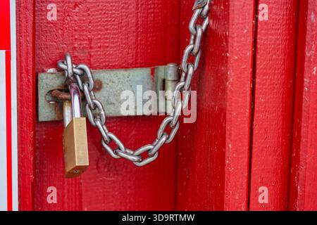 Flat wooden boards are painted deep red and a small padlock holds a short chain to the latch, the worn grain and tiny scuffs make the simple fastening Stock Photo