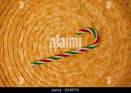 Red, green, and white striped candy cane ornament placed on a round woven straw mat, symbolizing Christmas, celebration, and cozy holiday decoration w Stock Photo