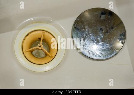 A bathroom sink shows the open drain with the shiny chrome cap lifted off and set nearby and the inner plastic trap is yellowed and full of wear, the Stock Photo