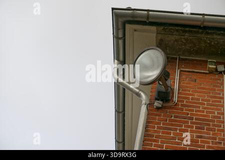 A close-up of a red clinker brick building corner with a wall-mounted large halogen floodlight, cables, and a rainwater downpipe against a cloudy sky. Stock Photo