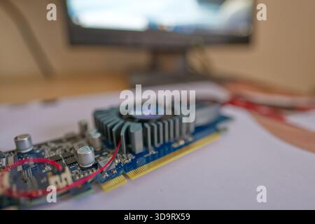 A close-up view of a computer graphics card on a desk with a blurred monitor in the background. The card features a cooling fan and various electronic Stock Photo