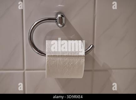 A close-up of a toilet paper roll on a chrome holder against a tiled wall. The roll is partially unrolled, showcasing its textured surface. Stock Photo