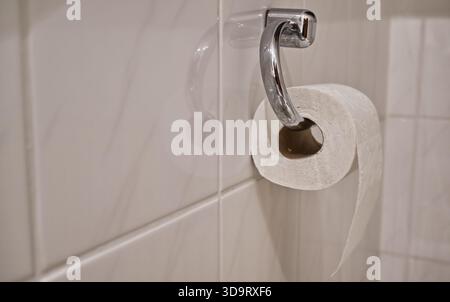 A close-up of a toilet paper roll hanging on a chrome holder against a tiled wall. The roll is partially unrolled, showcasing its soft texture and whi Stock Photo