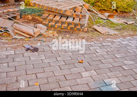 A construction site with stacked wooden pallets, scattered debris, and a paved pathway. A single glove lies on the ground among fallen leaves, indicat Stock Photo