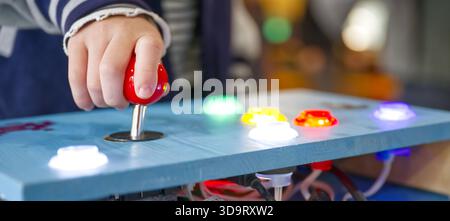A child's hand gripping a red joystick on a colorful control panel with illuminated buttons. The panel features various colored lights and a playful d Stock Photo