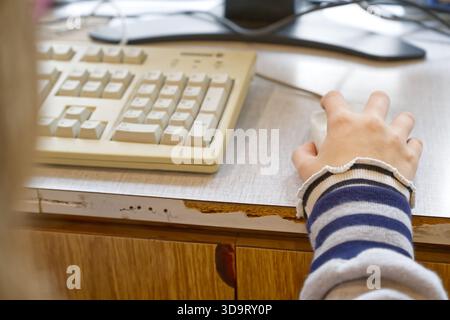 A close-up of a person's hand using a computer mouse next to a keyboard on a desk. The desk shows signs of wear, with a chipped edge, and the keyboard Stock Photo