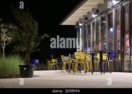 A modern outdoor seating area at night, featuring colorful yellow and blue chairs and tables. The setting is illuminated by soft lights, with greenery Stock Photo