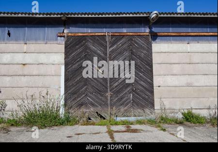 A weathered wooden door with a chevron pattern, set against a concrete wall. The surrounding area is overgrown with grass and weeds, under a clear blu Stock Photo