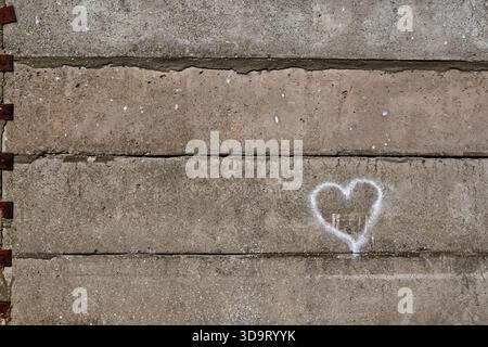 A textured concrete wall featuring a simple white heart shape drawn in chalk. The wall has a mix of gray and beige tones, with visible imperfections a Stock Photo
