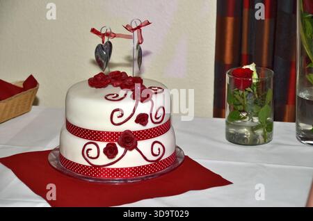 A beautifully decorated two-tier wedding cake with white fondant, red roses, and a red polka dot ribbon. The cake is topped with two silver heart-shap Stock Photo