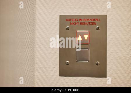 A close-up of an elevator control panel mounted on a wall. The panel has two buttons with arrows indicating up and down, and a warning message in Germ Stock Photo
