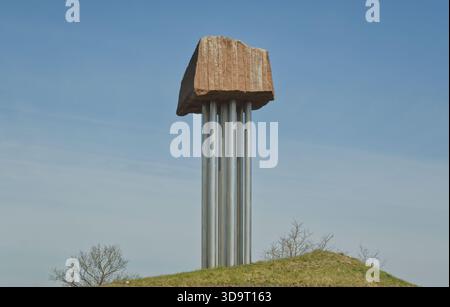 A large, abstract sculpture featuring a massive stone block balanced on several metallic pillars, set against a clear blue sky and grassy hill. Stock Photo