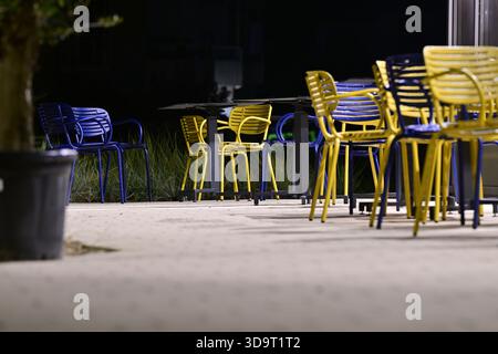A nighttime scene featuring colorful chairs in blue and yellow arranged around tables in an outdoor setting. The area is illuminated softly, creating Stock Photo
