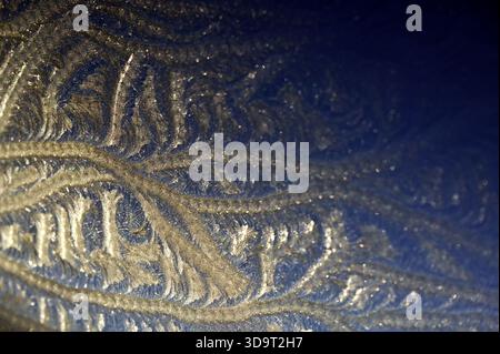 An abstract close-up shows complex patterns with natural textures and a mix of gold and blue tones, created by heavy frost on a skylight at sunrise. Stock Photo