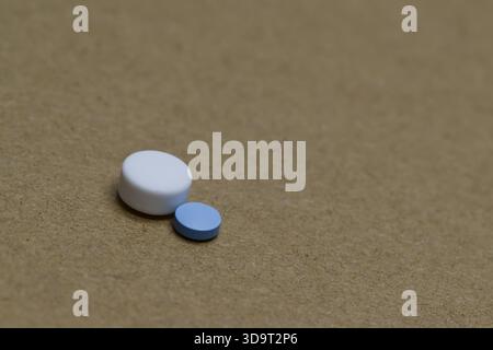 Two pills, one white and round, the other smaller and blue are placed on a textured brown surface. Stock Photo