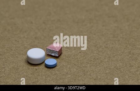 A close-up view of various pills on a textured eco brown surface. The image features a white round pill, a small blue pill, and a pink pill that is pa Stock Photo