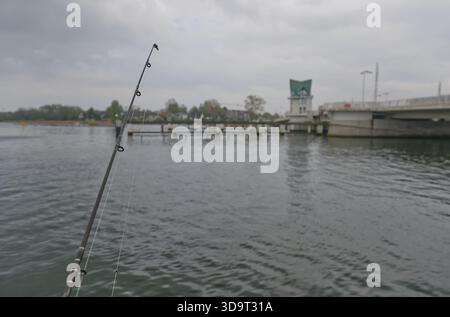 A fishing rod is positioned in the foreground, with a calm water surface reflecting the cloudy sky. In the background, a bridge and a structure resemb Stock Photo