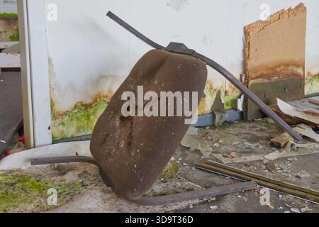An abandoned, damaged chair lying on its side in a dilapidated room. The floor is covered with debris and dirt, and the walls show signs of neglect wi Stock Photo